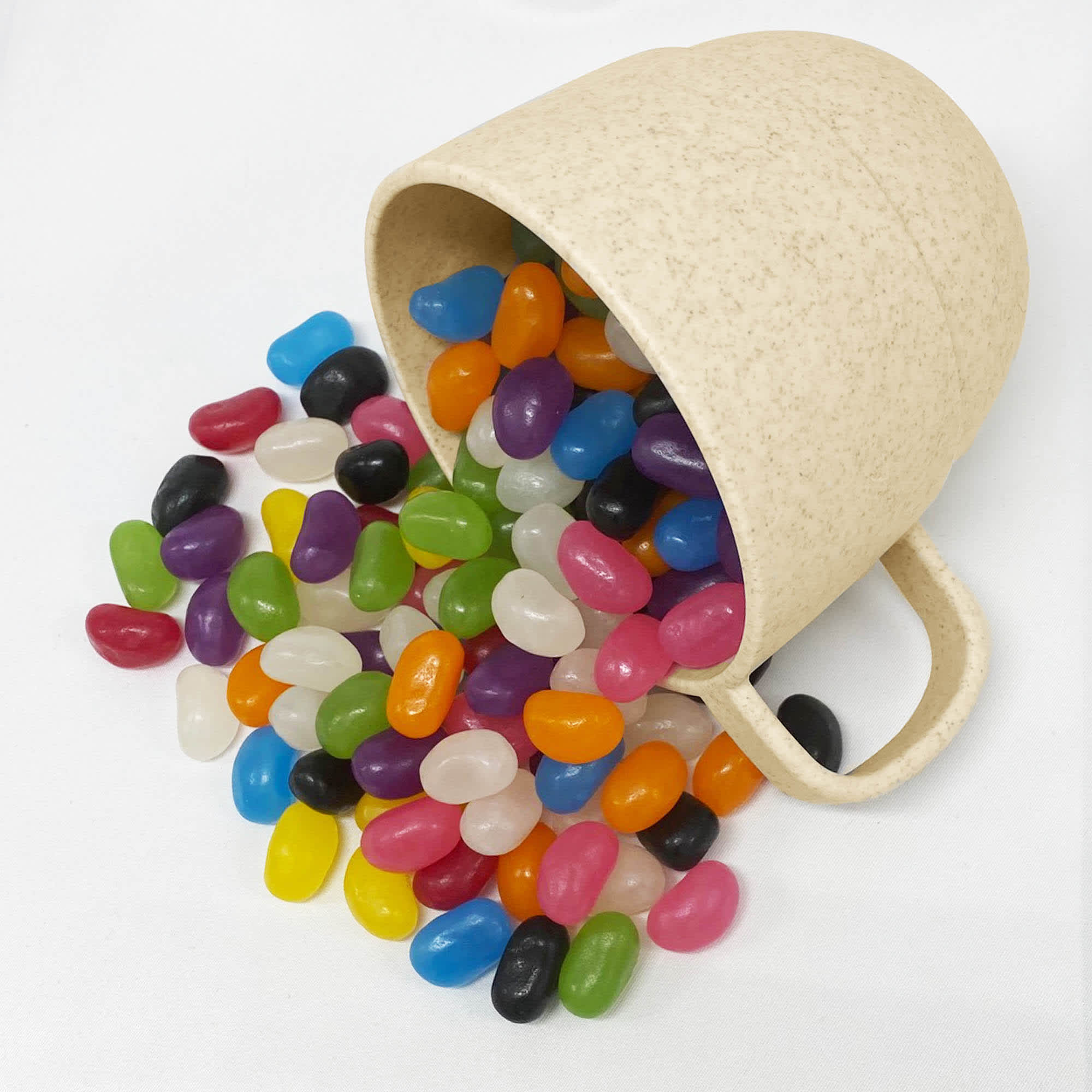 A cream wheat straw cup tipped over with assorted colourful jelly beans spilling out. The cup has a handle and a textured finish. Jelly Bean In Vetto Wheat Straw Cup - Image 1