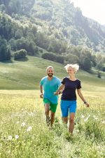 A couple running in a grassy field, both wearing bright blue and green trekking shorts. The landscape features lush green hills.