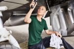 A green work t-shirt worn by a woman, seated on a mat near an aircraft's undercarriage, holding a tool.
