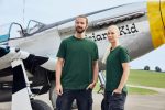 Two individuals wearing green BIO Stretch-T work t-shirts stand beside a vintage aircraft on a grassy field.