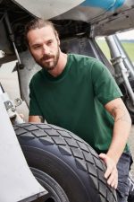 A man wearing a green BIO Stretch-T shirt adjusts a tyre near an aircraft. The shirt features a logo.