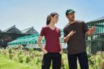 A maroon ladies' T-shirt and a brown men's T-shirt are displayed in a garden setting with greenhouses in the background.