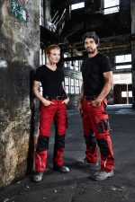 A pair of individuals in black t-shirts and red work trousers with black accents, standing in an industrial setting.