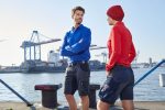 A pair of individuals wearing blue and red work shorts by a waterfront industrial area. The shorts feature pockets and branding.