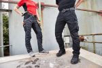 Two people wearing black stretch work pants, standing on a surface with industrial background, showcasing pockets and belts.