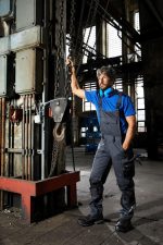 Workwear pants with bib in navy and grey, featuring a sturdy design and pockets, worn by a model in an industrial setting.