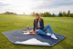 A picnic blanket in navy and white, laid out on grass with a woman sitting, surrounded by snacks and a drink bottle.