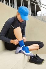 A person in a blue cap and black athletic wear adjusts their blue sports socks while seated on concrete steps.