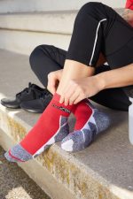 Sport socks in red with grey accents, featuring a logo, being worn by a person seated on a step.