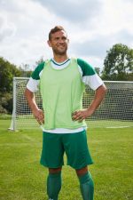 A young man stands confidently on a football pitch, wearing a bright green training bib, green shorts, and green socks.