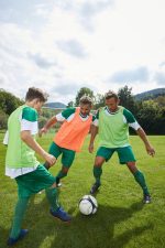 Three players in green and orange training vests engage with a soccer ball on a grassy field under a cloudy sky.