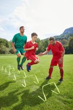 A training scene featuring two adult players in green and red shirts, coaching a young boy in a red shirt, jumping over hurdles.
