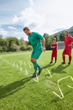 A man in green sports shorts and shirt performs agility drills over hurdles on a grass field, with two players in red watching.