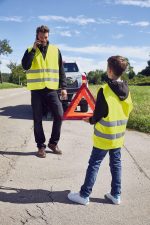 Two individuals wearing bright yellow safety vests stand by the roadside. The adult is on a phone, while the child holds a reflective triangle.