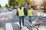 Two adults wear bright yellow safety vests while walking on a street, with a warning triangle nearby.