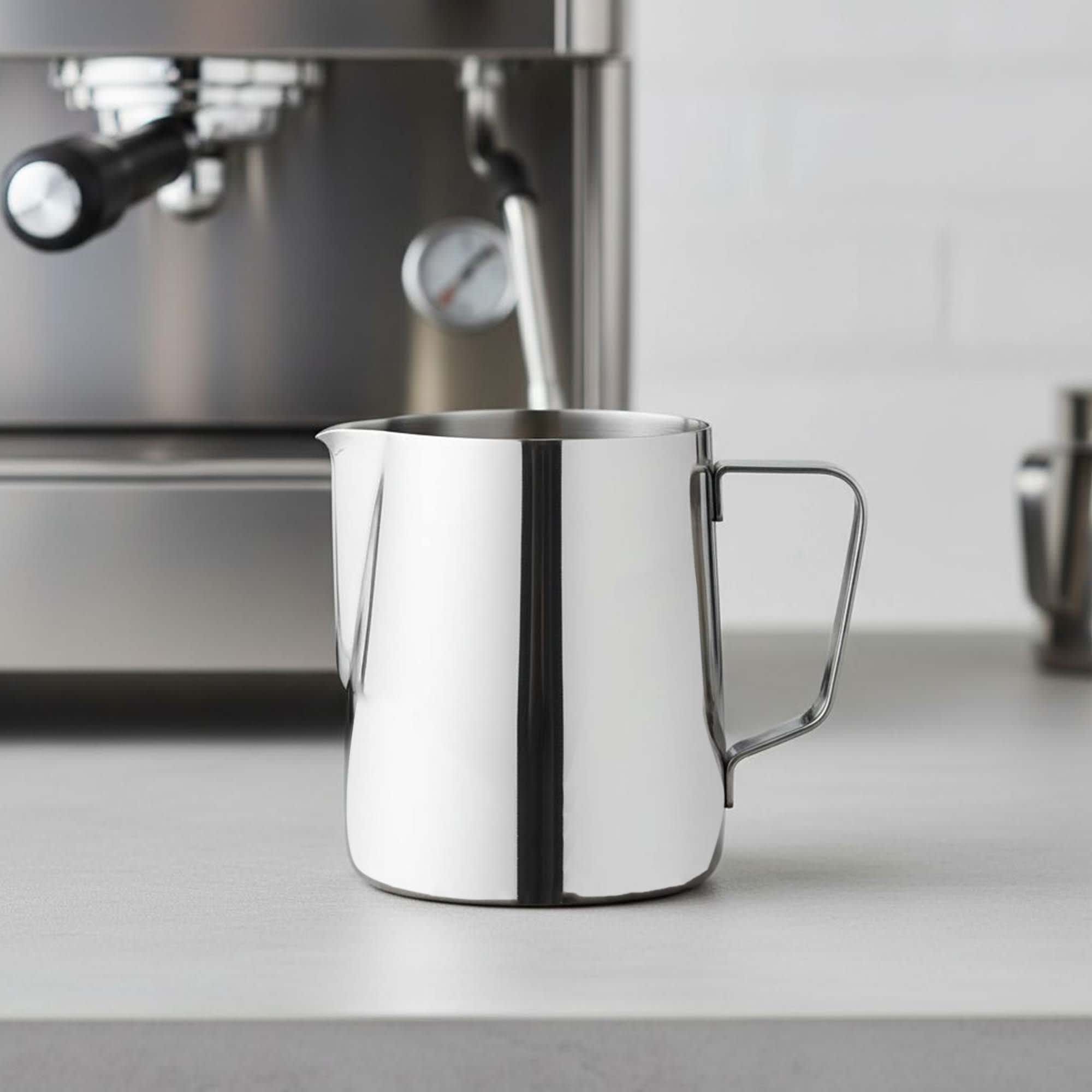 A stainless steel milk jug with a handle, positioned on a countertop near a coffee machine. Drop Milk Jug - Image 1