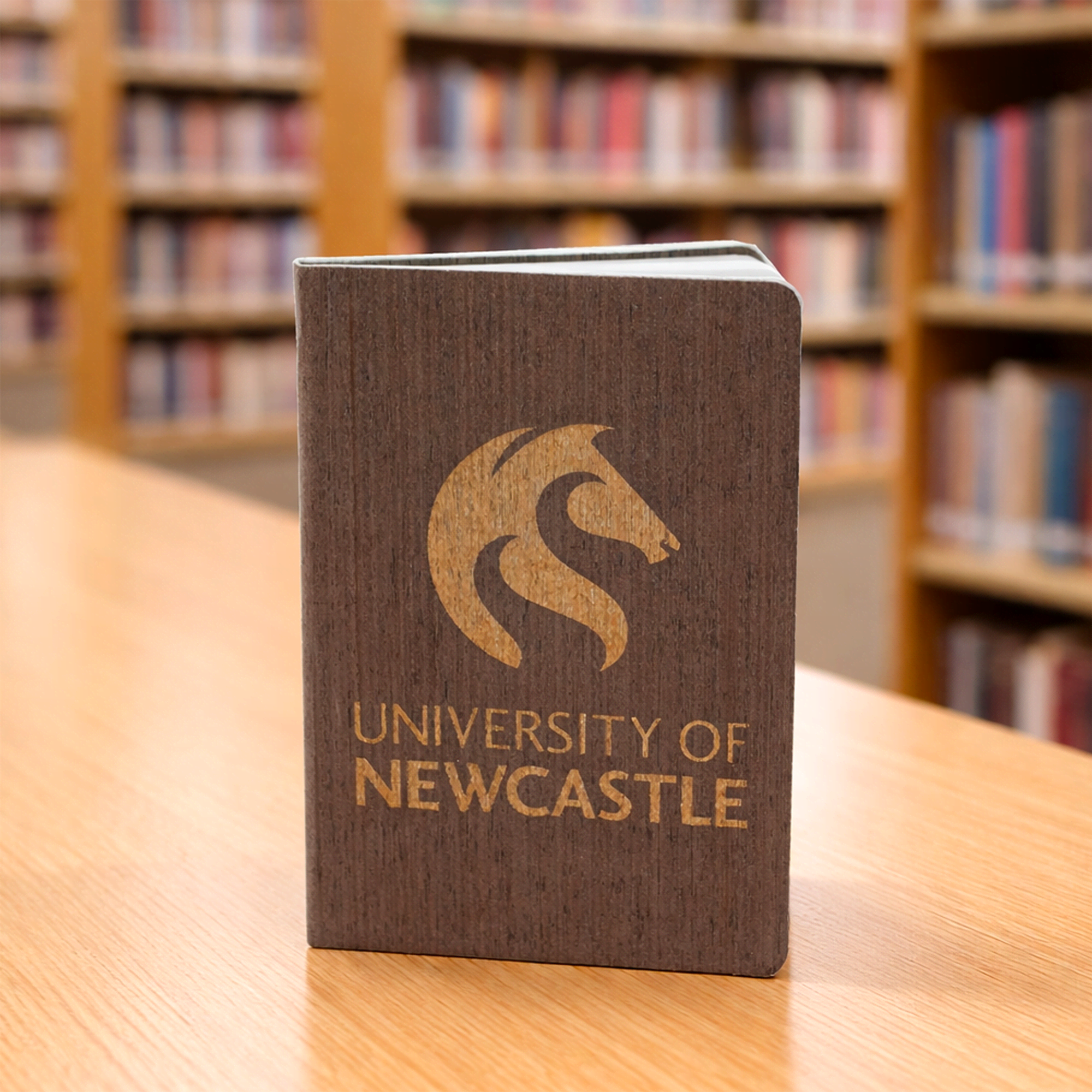 A brown eco notebook with a logo on the cover, placed on a wooden table in a library setting. The Barker Eco Notebook - B6 - Image 1