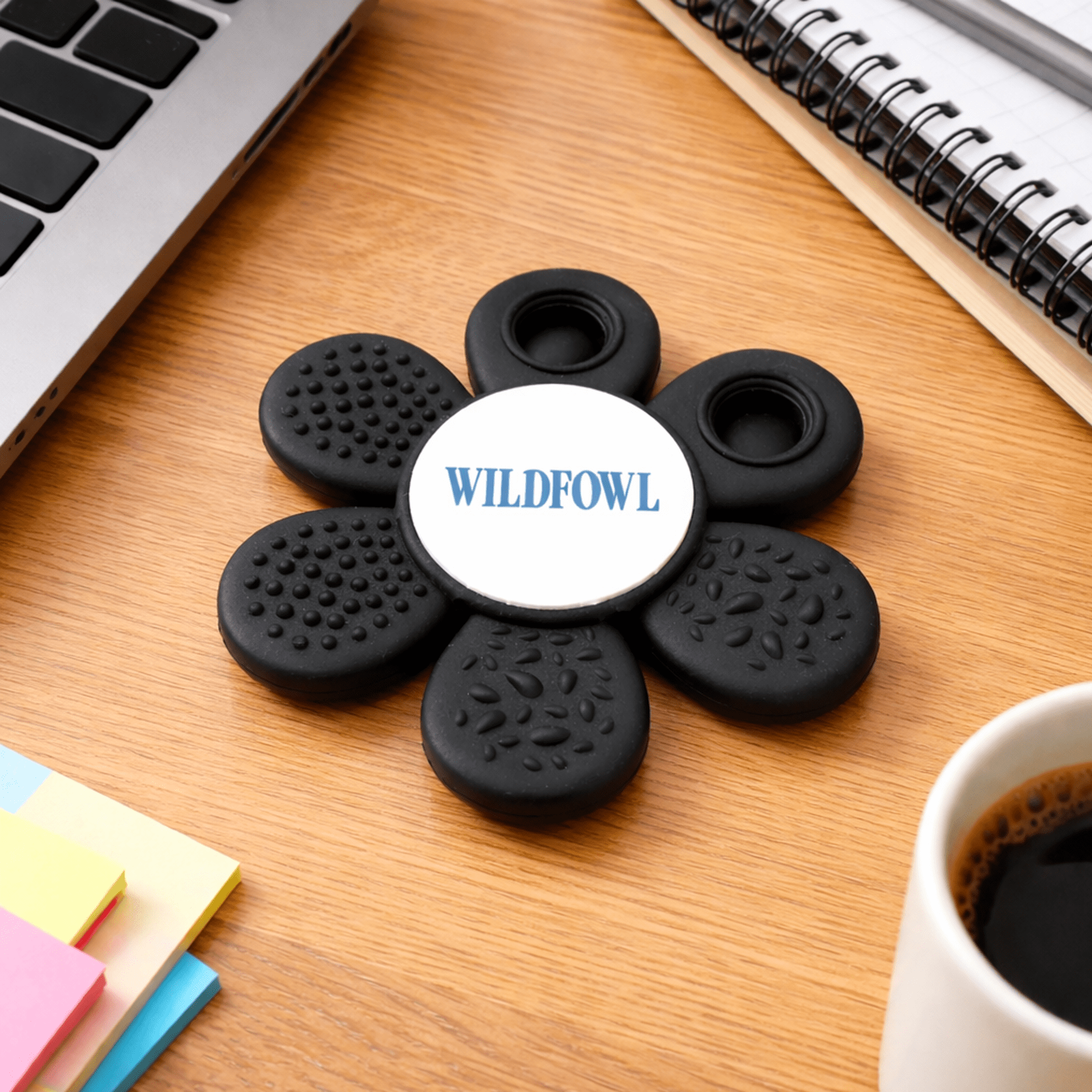 A black fidget spinner in a flower shape with a central logo, placed on a wooden desk near a coffee cup and stationery. The Spinzy - Image 1