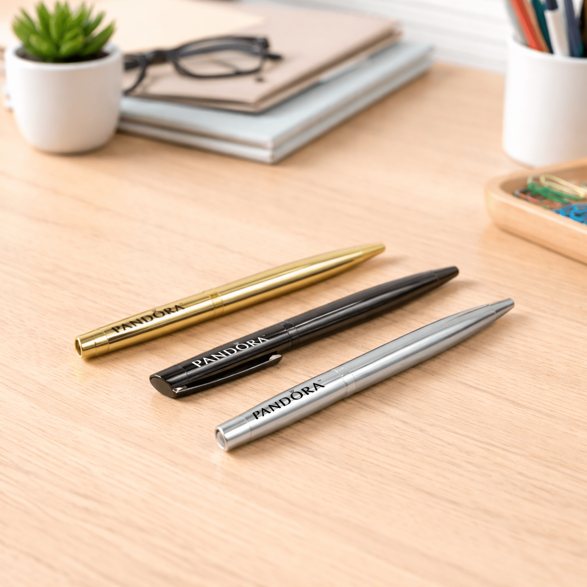 Three metal ballpoint pens in gold, black, and silver arranged on a wooden desk with stationery in the background. The Rhi Metal Ball Pen - Image 1