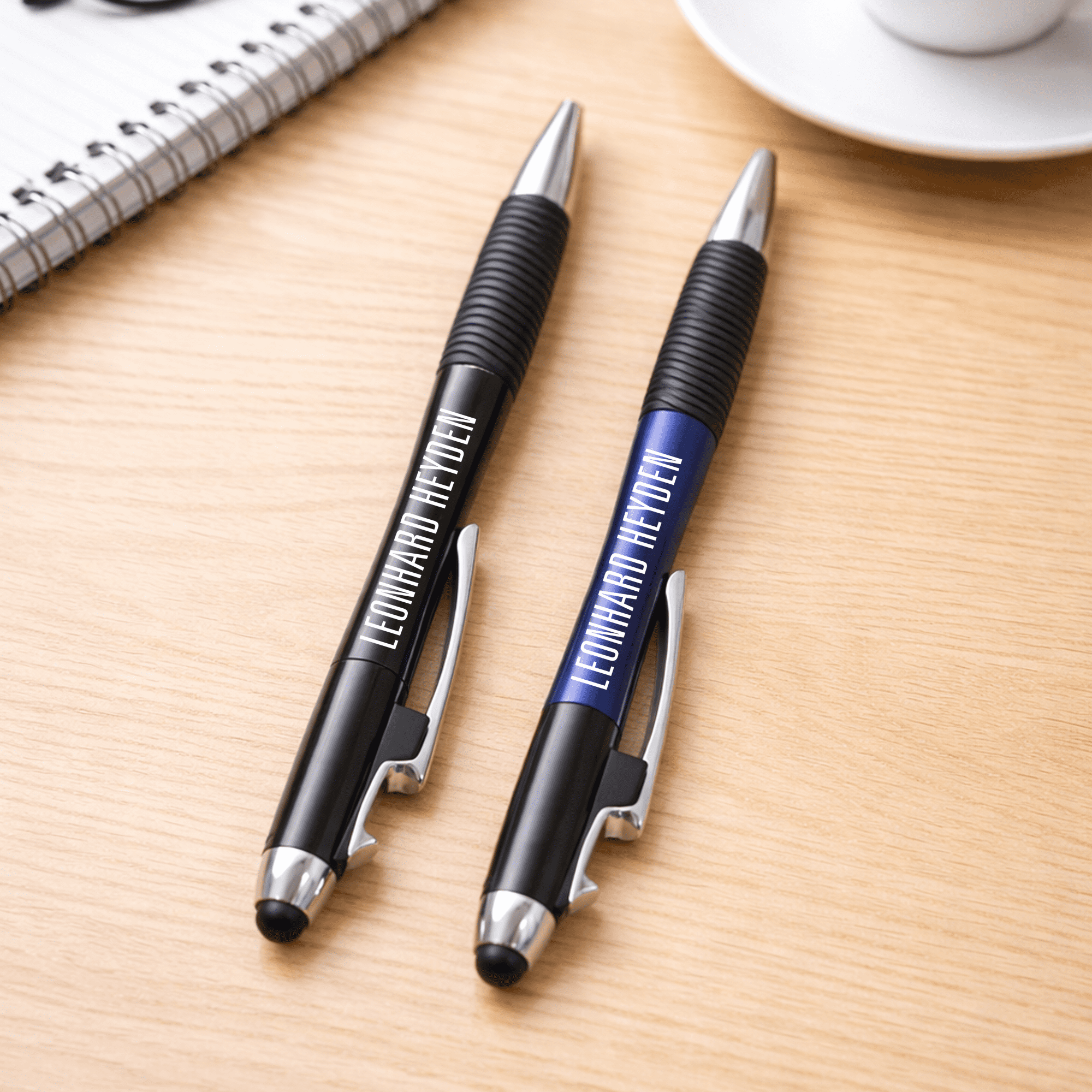 A pair of pens, one black and one blue, with grips, resting on a wooden surface near a cup and notebook. The Ironhide Pen - Image 1