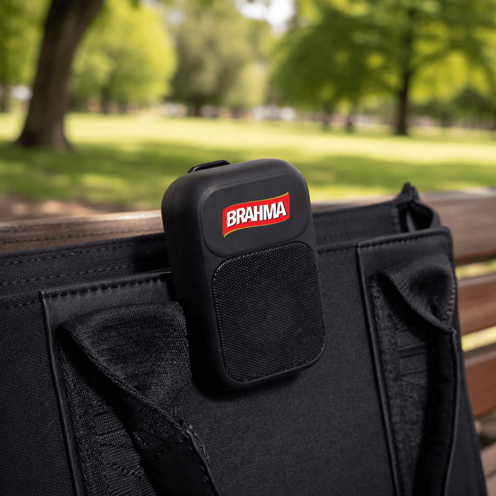 A black portable speaker with a logo, attached to a bag, set against a green park background. The Rebel Clip Speaker - Image 1