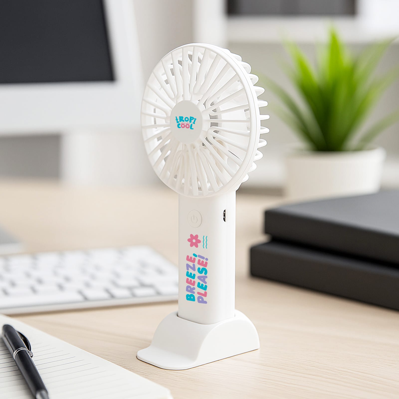 A white handheld fan with a base, featuring a logo and colourful text, placed on a desk near greenery and office supplies. Heatwave Fan - Image 1