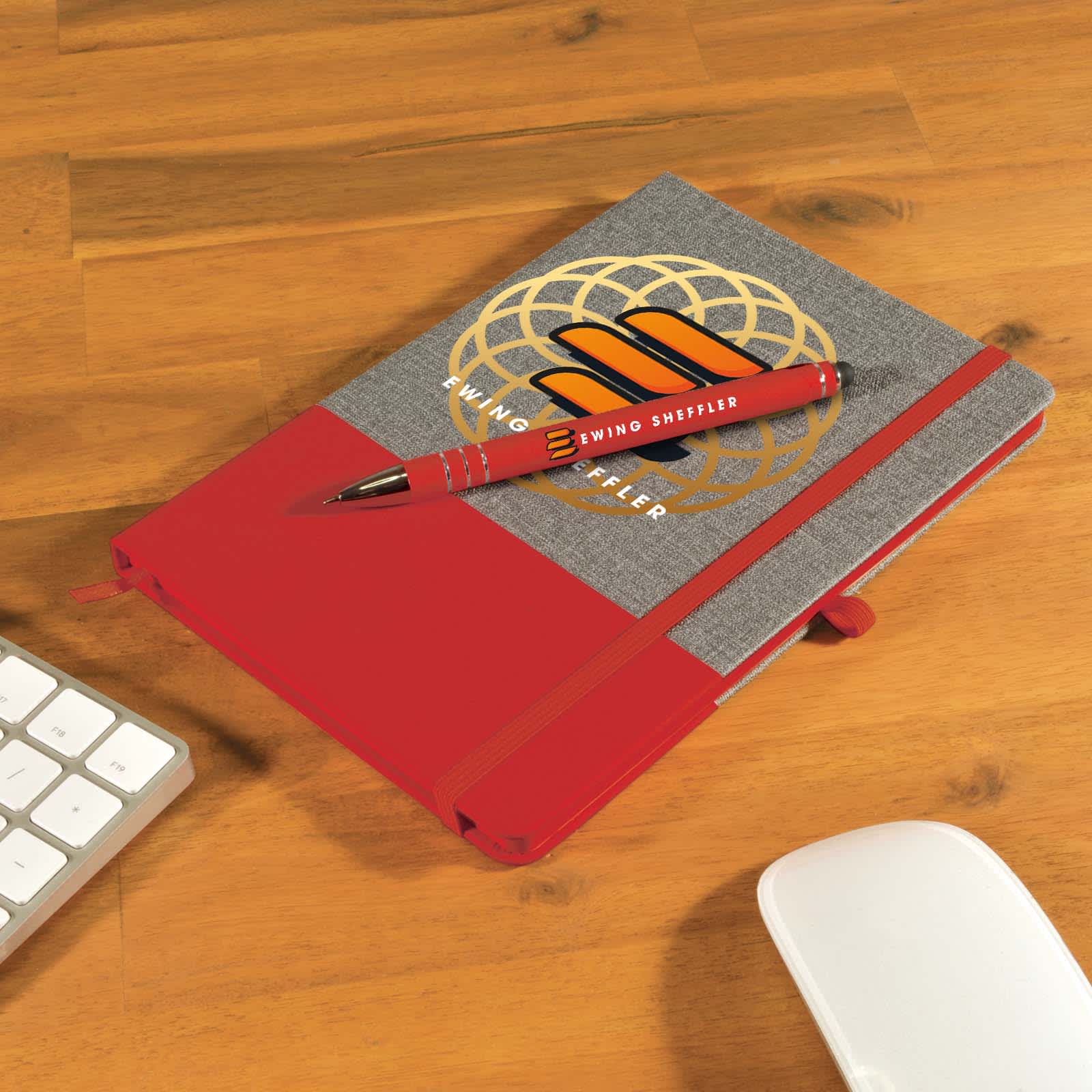 A grey notebook with a red band and a matching red pen placed on a wooden desk alongside a keyboard and mouse. Venture Bondi Notebook / Austin Pen - Image 1