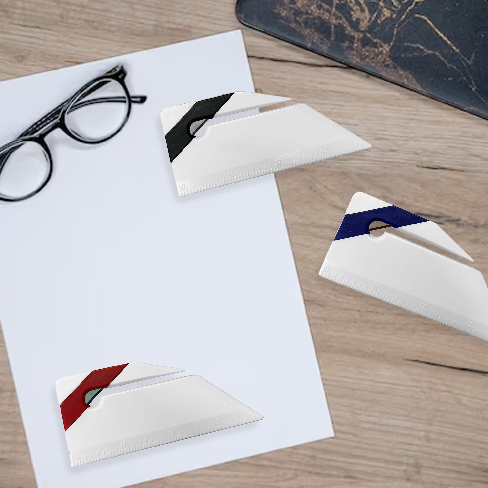 Three white letter openers in black, red, and blue, displayed on a wood surface with glasses and a sheet of paper. Ruler Letter Opener - Image 1