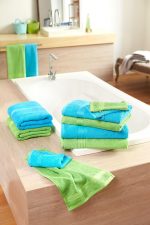 A stack of blue and green hand towels on a wooden surface near a bathtub, with more towels hanging nearby.