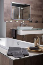 A bathroom scene featuring a white bathtub, grey towels, and a glass vase with decorative items on a wooden tray.