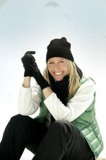 A woman wearing a black beanie and gloves, seated on snow, dressed in a green vest and black pants.