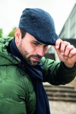 A man in a green jacket and navy scarf tips his blue cap, set against an urban backdrop.