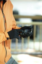 A person wearing black touch-screen knitted gloves uses a mobile device while dressed in an orange jacket.