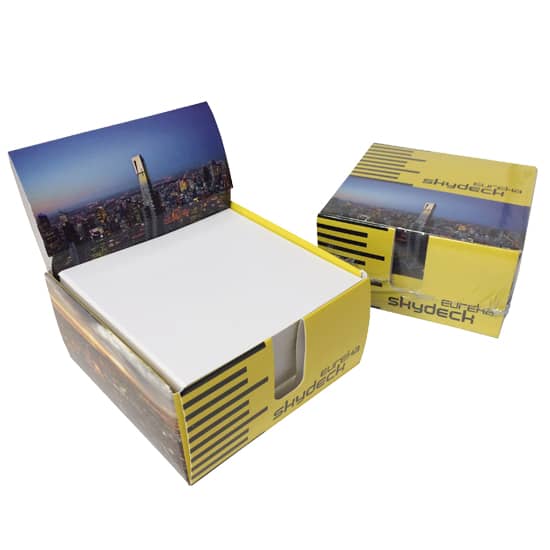 A card memobox with a scenic city skyline design, containing white memo papers. Predominantly yellow and featuring a logo. Memo paper in card memobox 1c (95x95x50mm) - Image 1