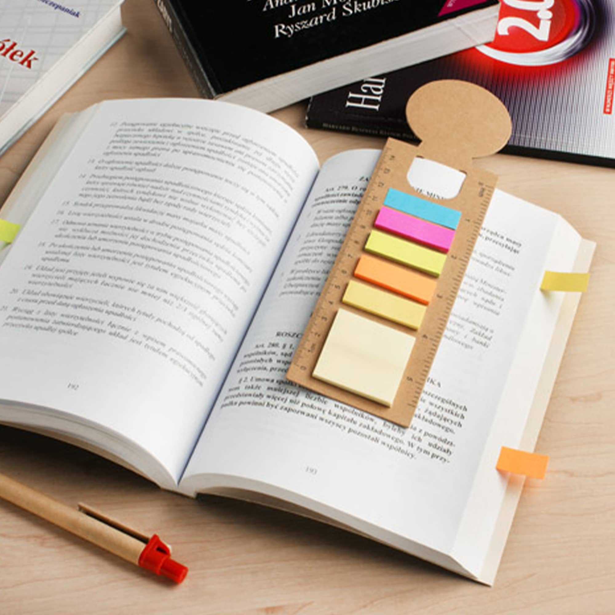 A bookmark ruler sticky note pad with various coloured sticky notes is placed on an open book, alongside a pen and books. Bookmark Ruler Sticky Note Pad - Image 1