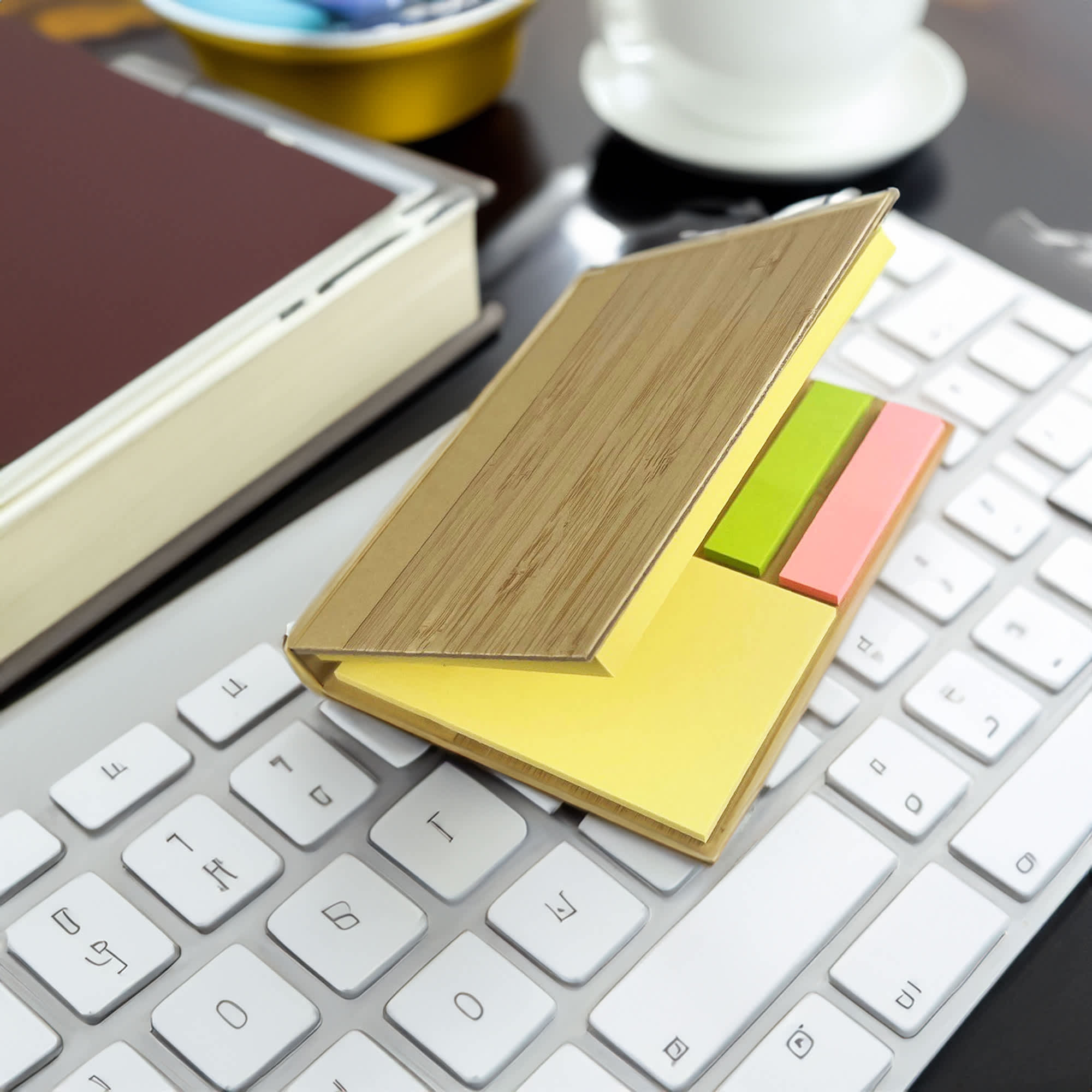 A bamboo sticky note holder with yellow, green, and pink sticky notes, placed on a white keyboard beside a book and a cup. Santo Bamboo Sticky Note - Image 1