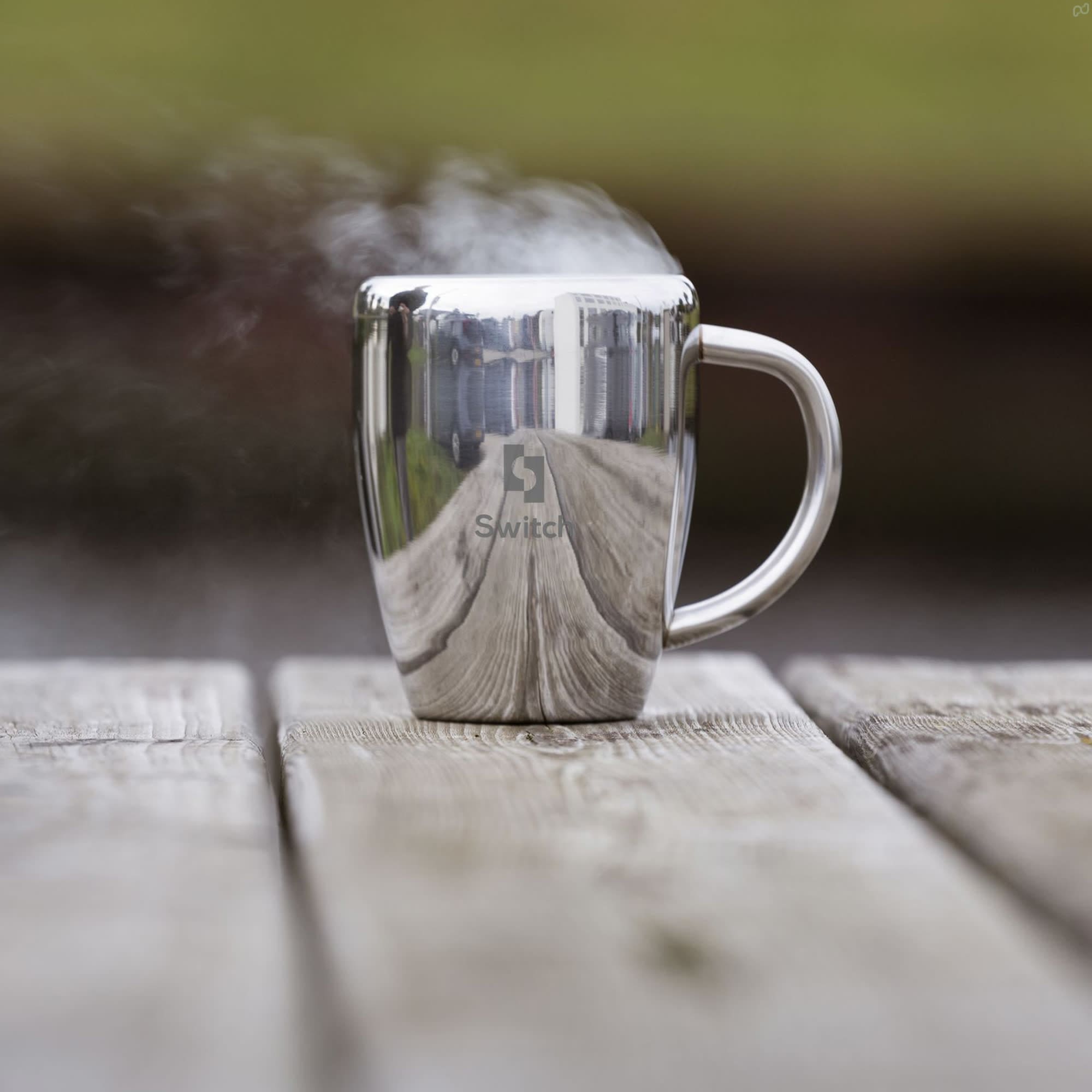 A silver coffee mug with a reflective surface and steam rising, placed on a wooden table. It features a logo. Compana Mug - Image 1