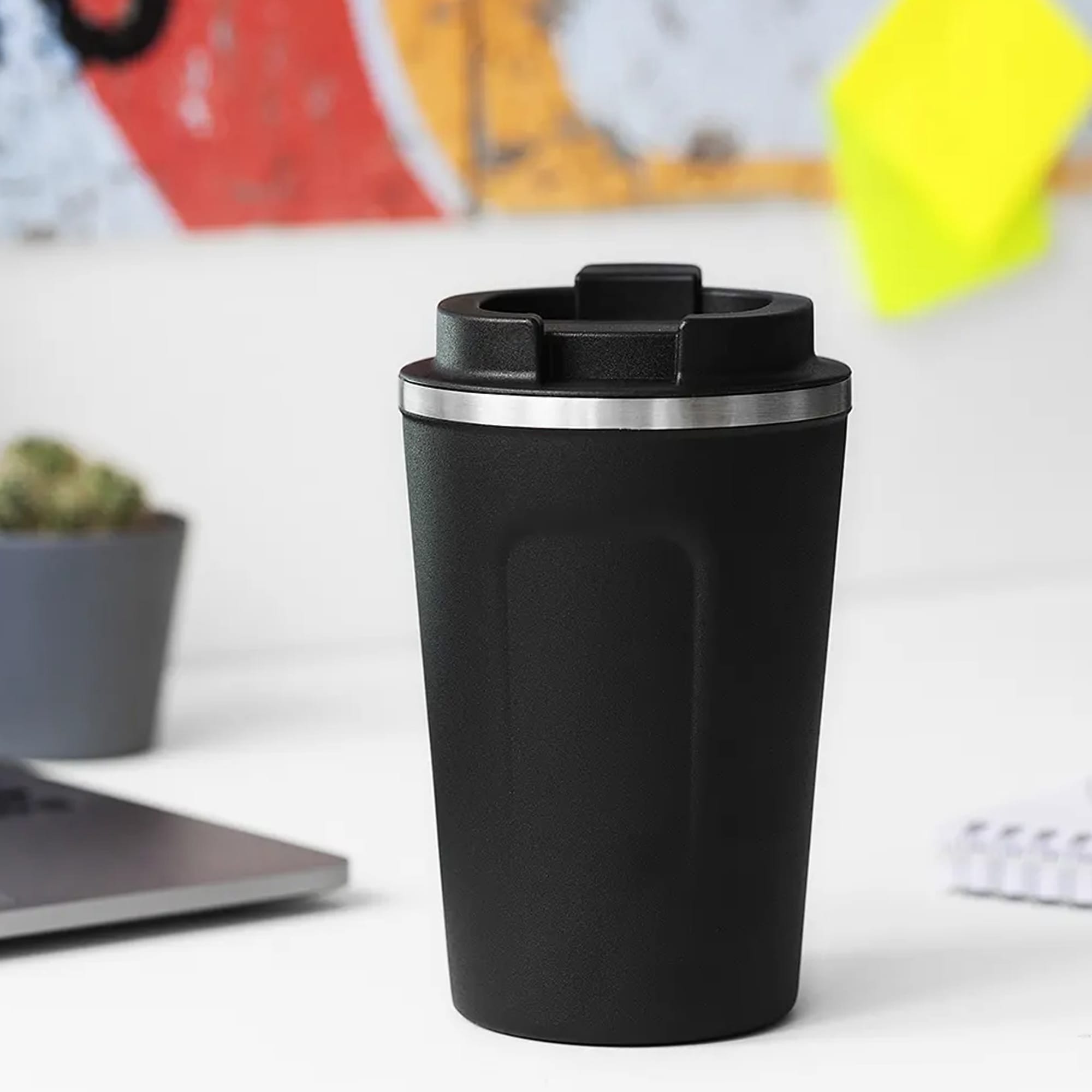 A reusable coffee cup in sleek black with a stainless steel lid, featuring a logo and set against a minimalist workspace. Kola Mug - Image 1
