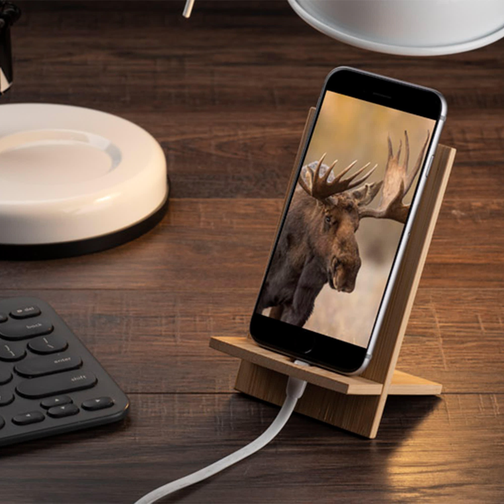 A bamboo phone holder displaying a phone with an image of a moose, set on a wooden desk beside a keyboard and lamp. Bamboo Phone Holder - Image 1