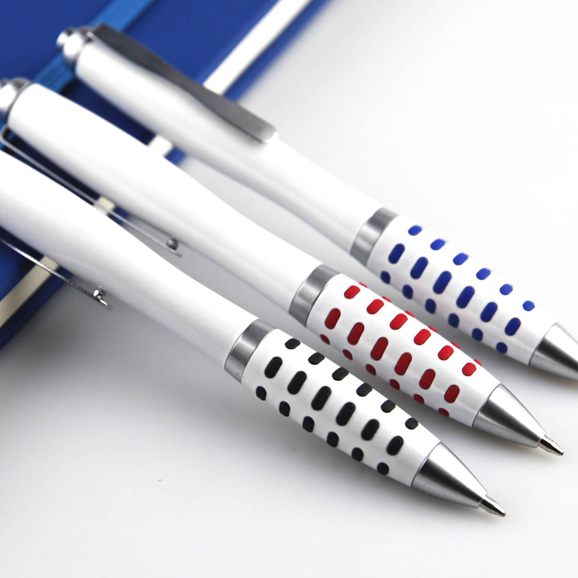 Three metal pens in white with patterned grips in red, blue, and black, resting on a notebook. Each pen features a logo. Aura Dot - Image 1