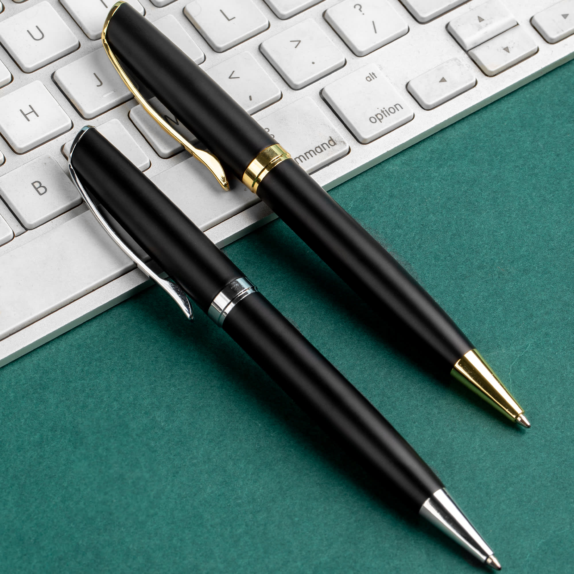 Two sleek black metal pens with silver and gold accents are placed next to a keyboard on a green surface. Prime - Image 1