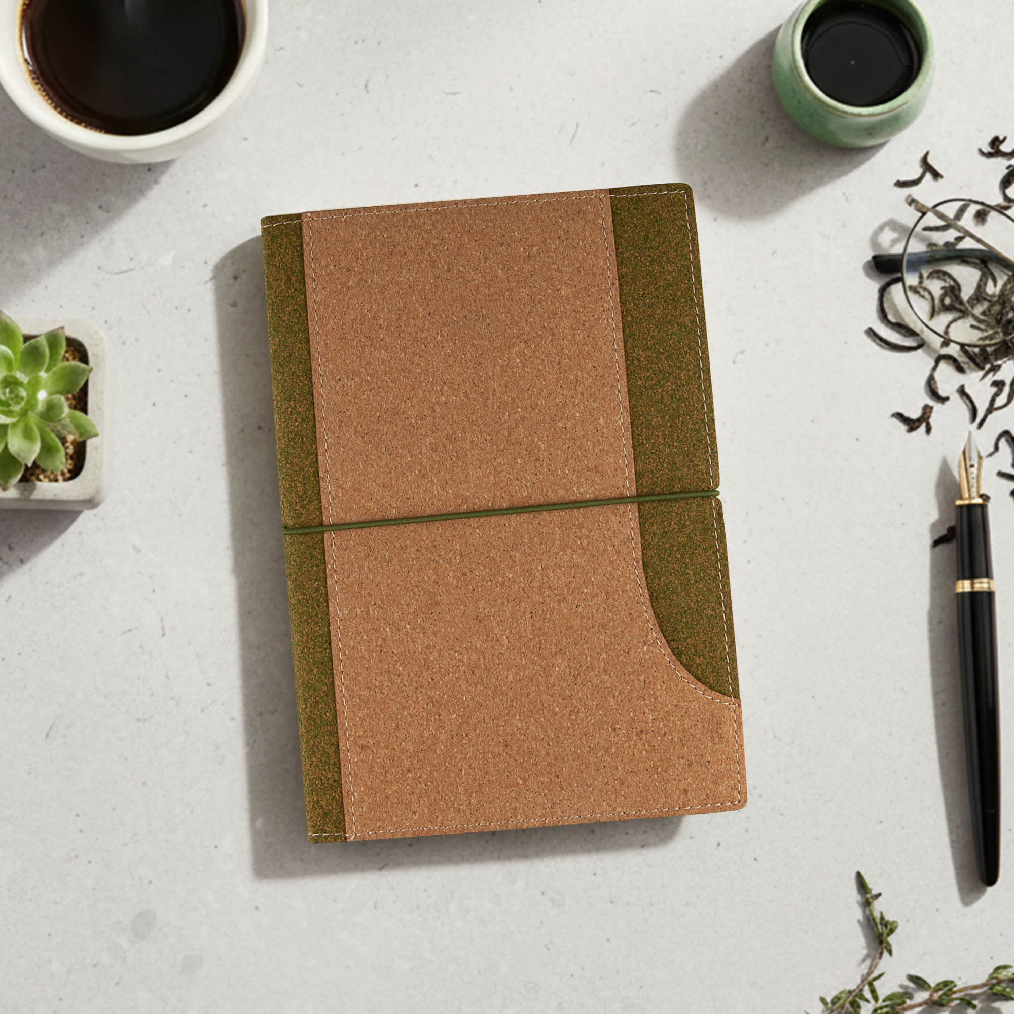 A cork notebook in brown and green, placed on a light surface with a cup, plant, and pen nearby. Tea Leaves and Cork Notebook - Image 1