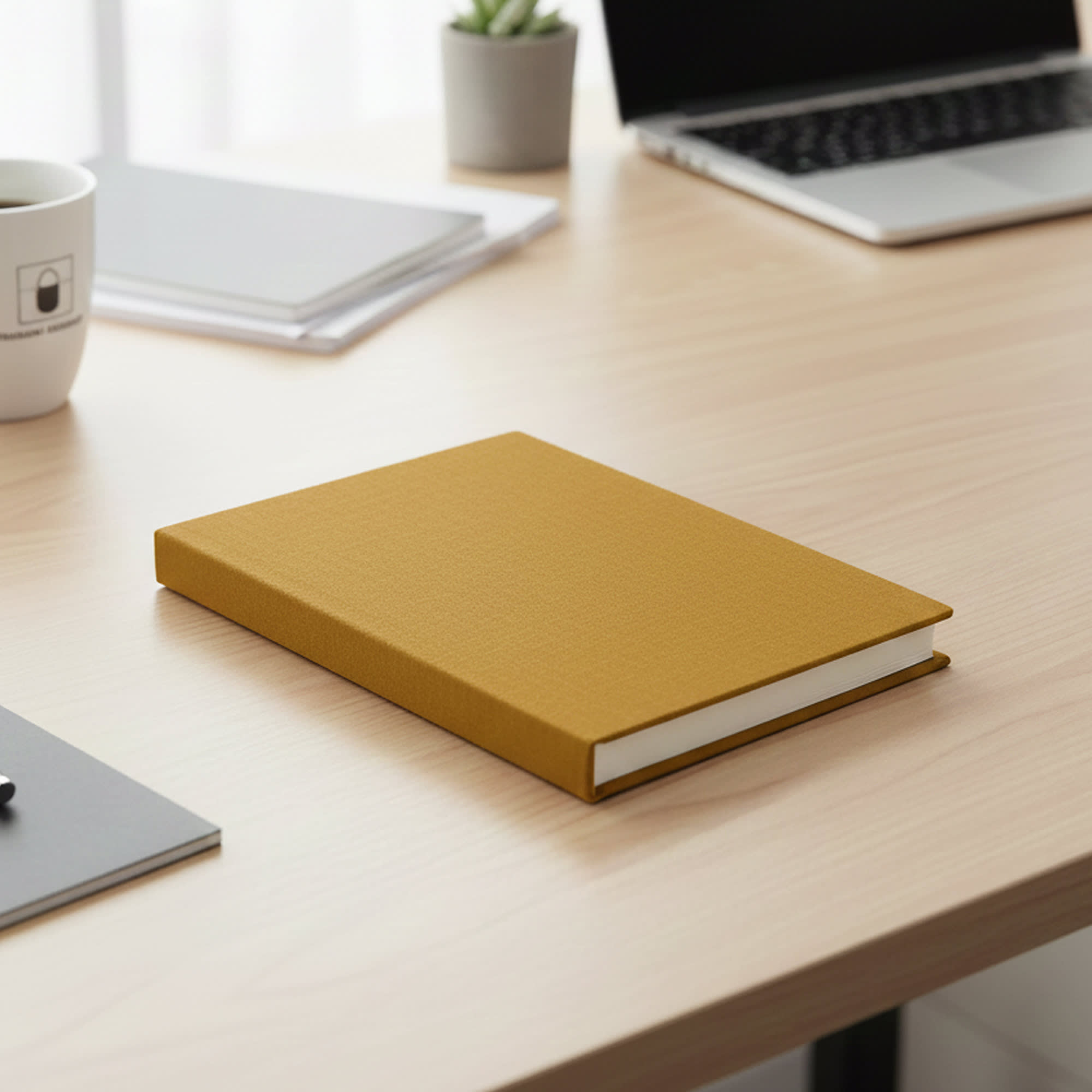 A mustard yellow A5 notebook with a textured cover resting on a wooden desk, alongside stationary and a laptop. Santiago Notebook A5 - Image 1