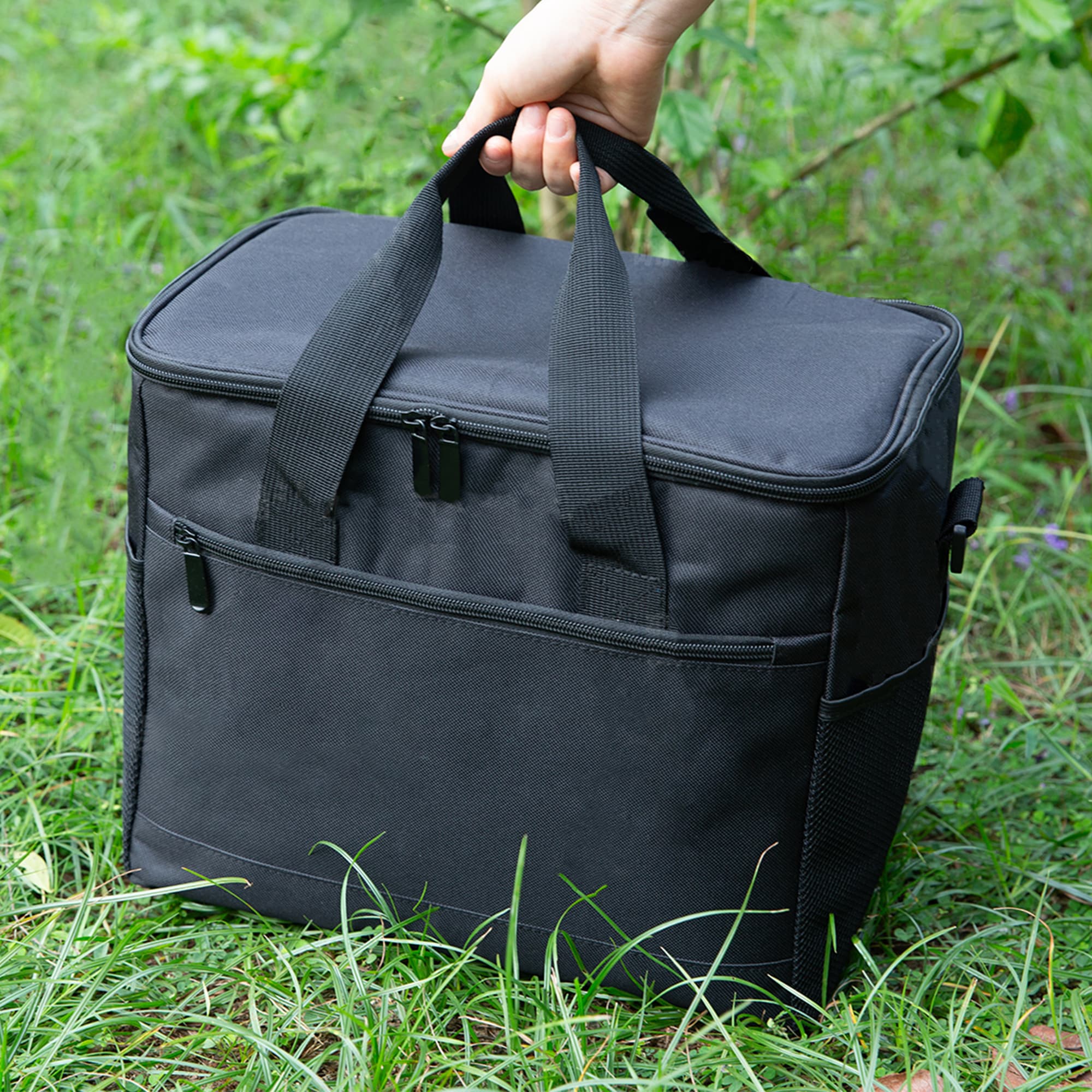 A black nylon cooler bag with carrying handles and a front pocket, positioned on green grass. Large Nylon Cooler Bag - Image 1