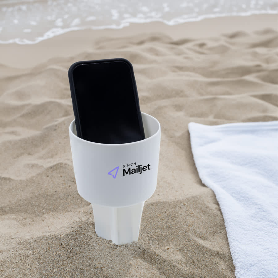 A white sand spike product holding a smartphone, placed on sand near a beach towel. The product features a branded logo. Sand Spike - Image 1