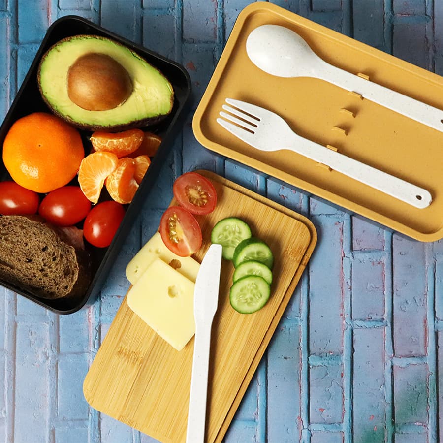 A two-layer lunch box in black and tan, featuring a variety of fresh ingredients and utensils on a wooden board. Two Layer Lunch Box - Image 1