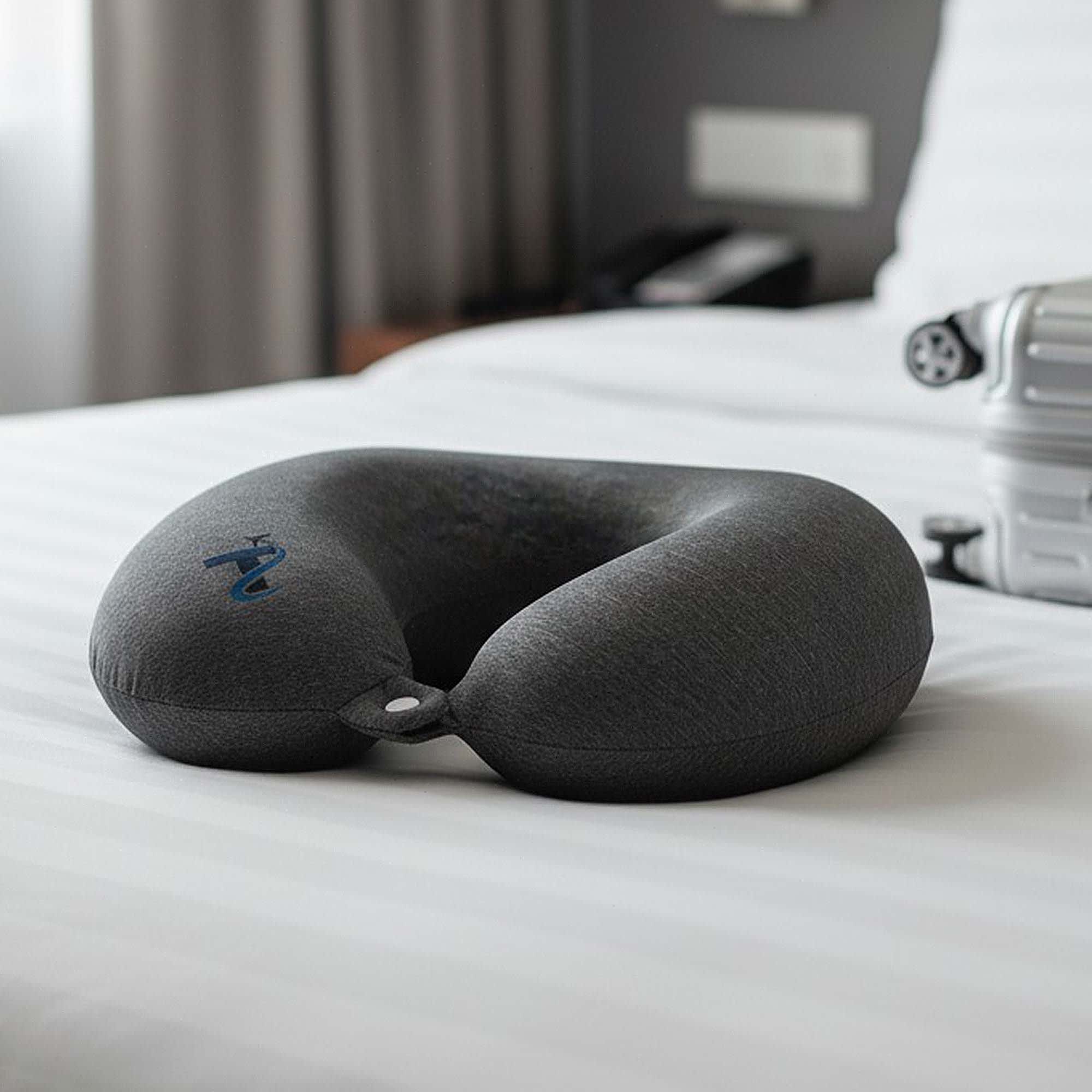 A grey neck pillow with a U-shape design rests on a neatly made bed. It has a small loop for attachment. Dona Memory Neck Pillow - Image 1