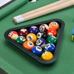 A triangle rack filled with colourful billiard balls on a green felt surface, with two cue sticks nearby.
