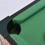 A close-up view of a mini billiard game's green felt surface and pocket, with a black frame. It features a logo.