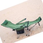 A green camping lounge chair with adjustable armrests and a headrest, resting on sandy ground.